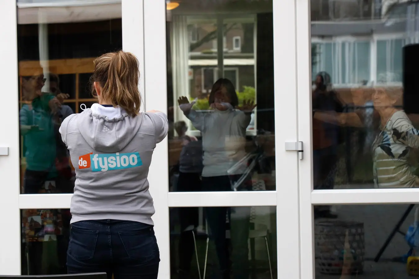 De fysiotherapeut blijft buiten achter het raam en geeft gymles aan de bewoners van Het Gofferthuis