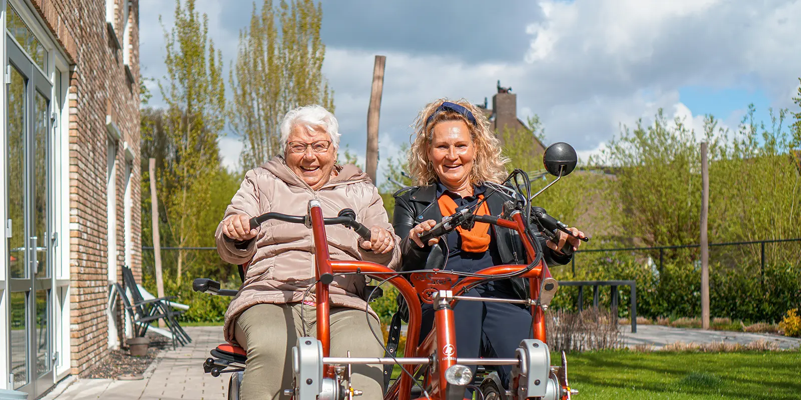 Een bewoonster van het Abtswoudehuis fietst samen met haar dochter op de duofiets door de tuin.