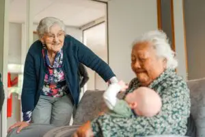 Een zorgmedewerkter is met haar pasgeboren baby op bezoek in Het Prins Claushuis. Een bewoonster heeft haar op de arm terwijl een andere bewoonster eroverheen gebogen staat en vertederd toekijkt.
