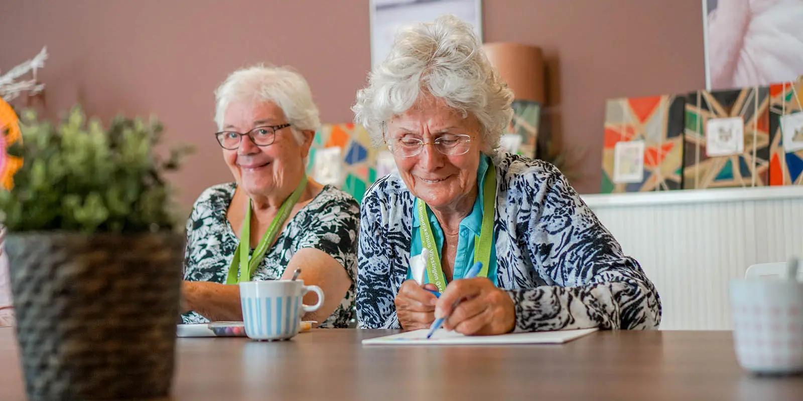 Twee bewoners met dementie schrijven iets op een papier. Ter illustratie bij een blog over gedichten over de feestdagen, geschreven door bewoners van een van de kleinschalige woonzorglocaties voor mensen met dementie van Dagelijks Leven.
