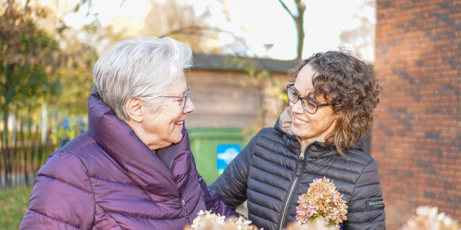 Een bewoner wandelt samen met haar dochter door de tuin van een woonzorglocatie van Dagelijks Leven voor mensen met dementie. Ze zorgt graag voor haar medebewoners en voelt zich inmiddels helemaal thuis.