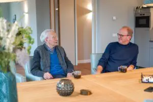 Bewoner van Het Blaauwhofhuis in Joure, kleinschalig wonen voor mensen met dementie in Friesland, zit met zijn zoon aan de koffie aan de eettafel in de woonkamer. Ze kijken lachend naar elkaar.