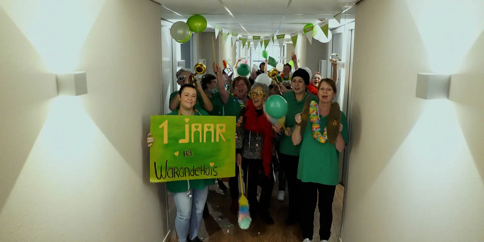 Bewoners en medewerkers van Het Warandehuis, kleinschalige woonzorglocatie voor mensen met dementie, samen dansend op de gang, met feestelijke hoedjes, zonnebrillen en muziekinstrumenten.