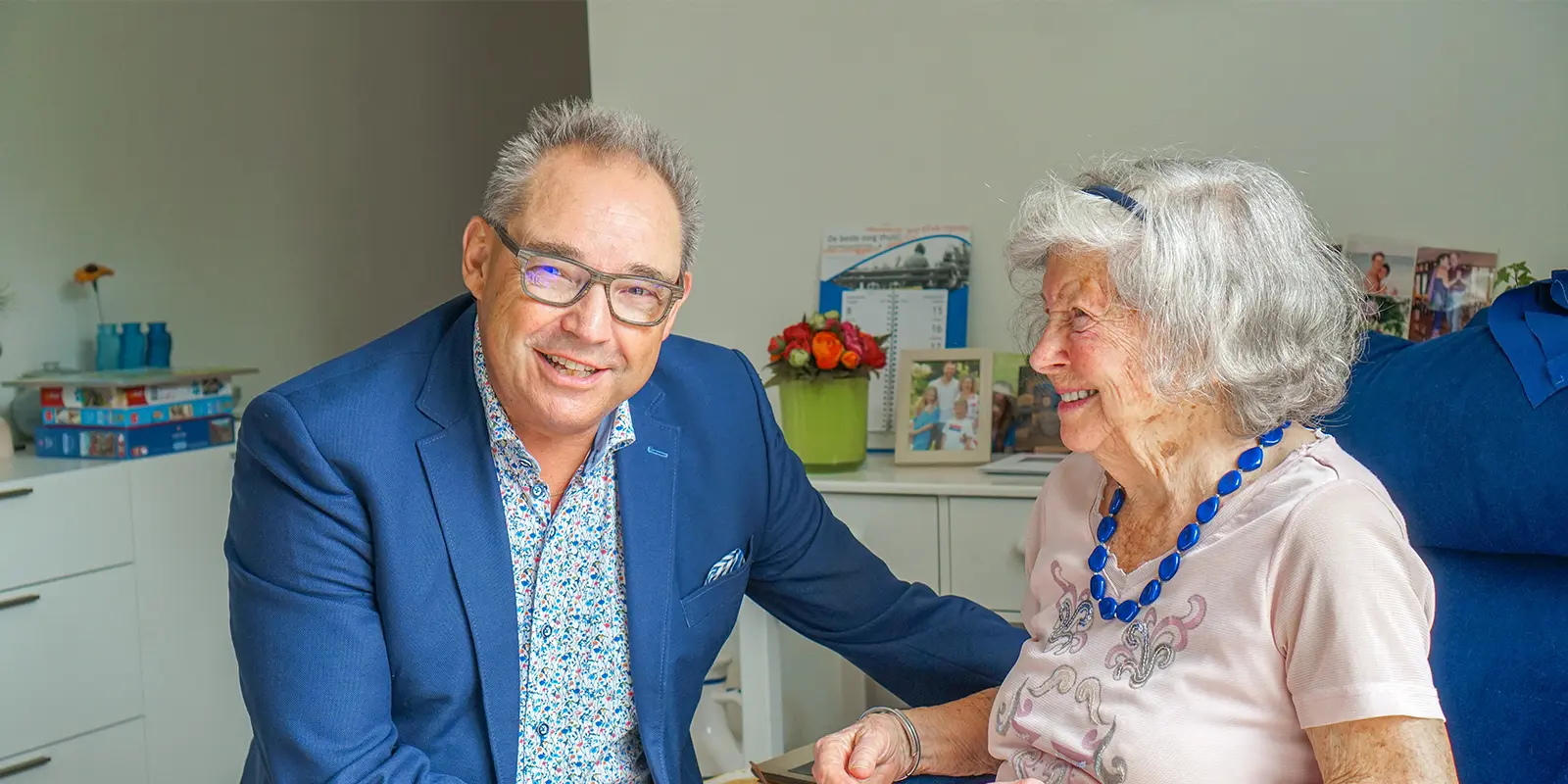 Specialist ouderengeneeskunde zit bij een bewoonster. De SO lacht in de camera, de bewoonster van de zorglocatie van Dagelijks Leven, voor mensen met dementie, lacht naar de dokter.