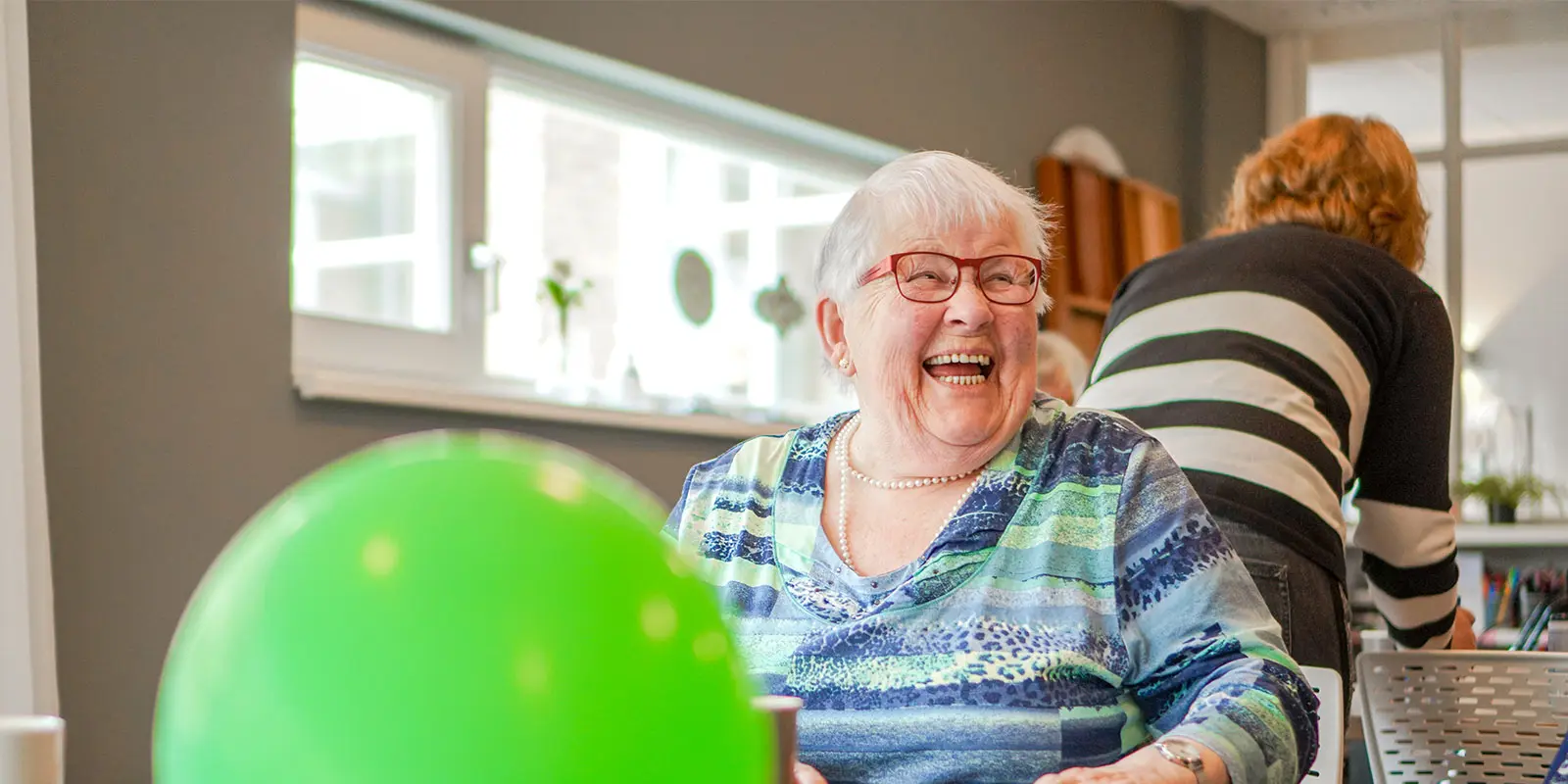Humor houdt je jong, ook als je eigenlijk al wat ouder bent, weet de activiteitenbegeleider van een huis van Dagelijks Leven voor mensen met dementie. Op de foot zien we een bewoonster heel hard lachen. Ze zit aan tafel, voor haar een groene ballon.