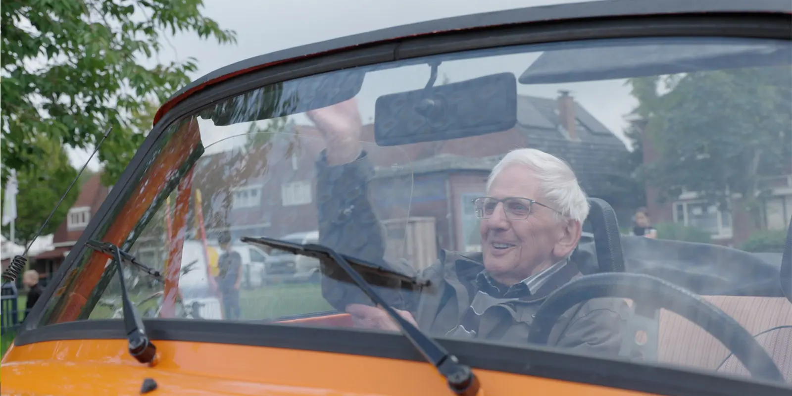 Bewoner met dementie zit in een Trabant op de passagiersstoel. Hij lacht en zwaait uit het raam. Hartenwens.