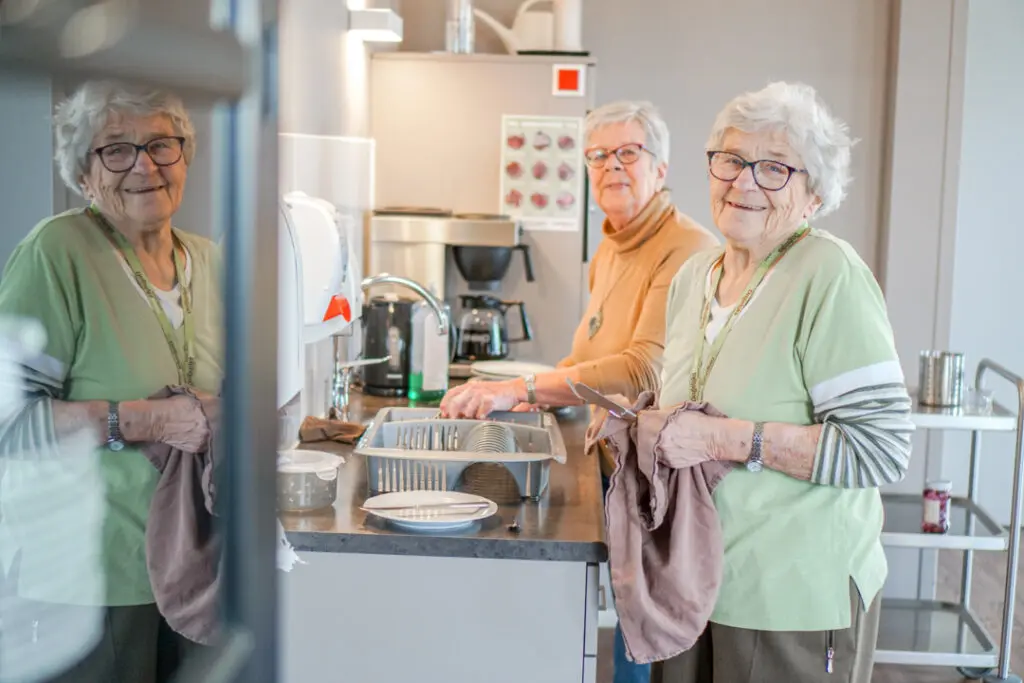 Twee bewoonsters met alzheimer van de kleinschalige zorglocatie doen samen de afwas en lachen naar de camera. De bewoonsters wonen in het verzorgingshuis en helpen graag met allerlei huishoudelijke klusjes