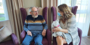 Verzorgende IG Isabel zit naast een bewoner van Dagelijks Leven in een fauteuil. Ze laat hem een knuffelhond zien.