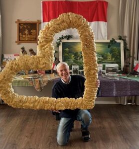 Bewoner met dementie heeft als hartenwens nog eens terug te keren naar zijn geboorteland Indonesië. Dagelijks Leven liet deze wens op zijn 79e verjaardag uitkomen met een echte Indonesische themadag. We zien hier de bewoner voor een ingerichte thematafel terwijl hij lachend door een groot gouden hart kijkt. 