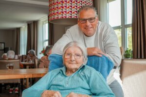 Bewoonster Abtswoudehuis zit in een rolstoel in de huiskamer van Dagelijks Leven, haar zoon staat achter haar en leunt op de stoel. Ze lachen naar de camera.