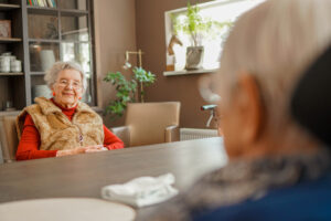 Twee bewoonsters zitten tegenover elkaar aan tafel in een woonzorglocatie van Dagelijks Leven. Een zien we op de rug, de ander lacht in de camera. Dagelijks Leven bouwt twee huizen voor mensen met dementie in Sittard.