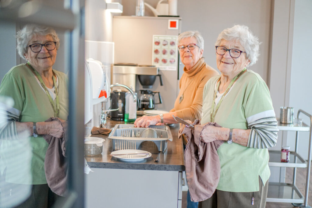 Twee bewoonsters met alzheimer van de kleinschalige zorglocatie doen samen de afwas en lachen naar de camera. De bewoonsters wonen in het verzorgingshuis en helpen graag met allerlei huishoudelijke klusjes