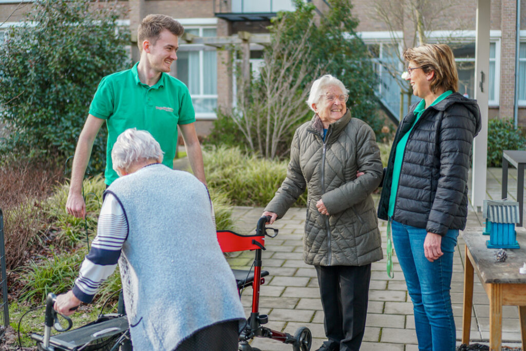 Twee bewoonsters, een met alzheimer en eentje met vasculaire dementie lopen samen met twee zorgmedewerkers een rondje om de zorglocatie voor mensen met dementie van Dagelijks Leven